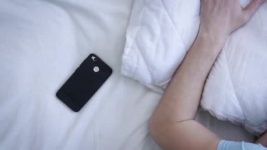 Genç adam uyandı, telefona baktı ve tekrar uyumaya başladı. Telefondaki çalar saat. Yorgun bir adam yatak odasında yatakta uyanıyor. Modern teknoloji kavramı