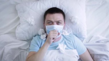 Tıbbi maskeli bir adam yatakta grip yüzünden öksürüyor. Boğazı ağrıyan tıbbi maskeli hasta bir adam. Coronavirus Covid-19. Sağlık kavramı