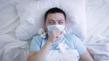 Tıbbi maskeli bir adam yatakta grip yüzünden öksürüyor. Boğazı ağrıyan tıbbi maskeli hasta bir adam. Coronavirus Covid-19. Sağlık kavramı
