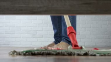 Genç adam oturma odasında paspasla yatağın altını temizliyor.