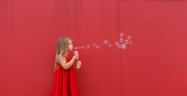 Ragazza bionda bambino in abito rosso su uno sfondo del muro soffia bolle . — Foto Stock