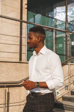 Beyaz gömlekli ve klasik pantolonlu yakışıklı Afrikalı bir adam dışarıda yürüyor. Yaşam tarzı portresi