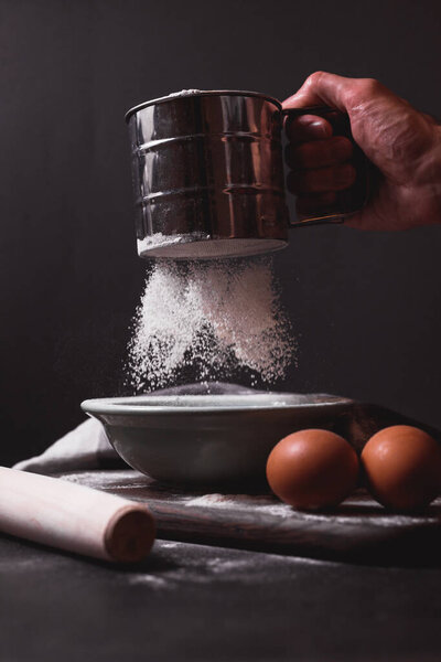 Рука держит металлический просеиватель муки, решето, посыпанное мукой. Просеиваю муку. Подготовка к выпечке. Низкий ключ фото