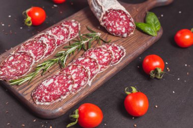 Koyu ahşap tahtada füme jambon ve biberiye ve kiraz domatesli dilimler.