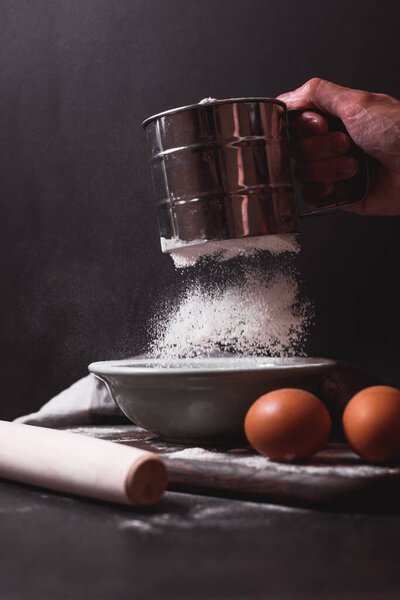 Рука держит металлический просеиватель муки, решето, посыпанное мукой. Просеиваю муку. Подготовка к выпечке. Низкий ключ фото