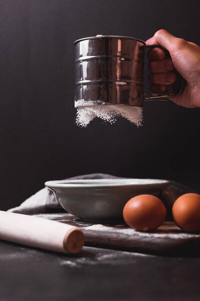 Рука держит металлический просеиватель муки, решето, посыпанное мукой. Просеиваю муку. Подготовка к выпечке. Низкий ключ фото.