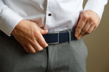 Mavi pantolonlu ve beyaz gömlekli bir adam kahverengi deri pantolon kemerini ilikledi. Elinde bir saat var..