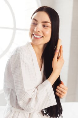 Genç esmer kadın, sağlıklı ve parlak saçlarını tarıyor beyaz bornozla küvette oturuyor.