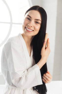 Genç esmer kadın, sağlıklı ve parlak saçlarını tarıyor beyaz bornozla küvette oturuyor.