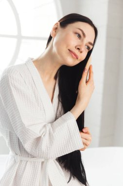 Genç esmer kadın, sağlıklı ve parlak saçlarını tarıyor beyaz bornozla küvette oturuyor.