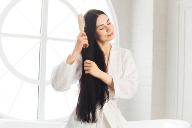 Genç esmer kadın, sağlıklı ve parlak saçlarını tarıyor beyaz bornozla küvette oturuyor.