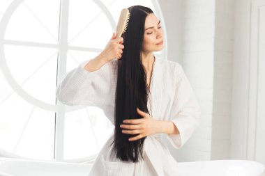 Genç esmer kadın, sağlıklı ve parlak saçlarını tarıyor beyaz bornozla küvette oturuyor.