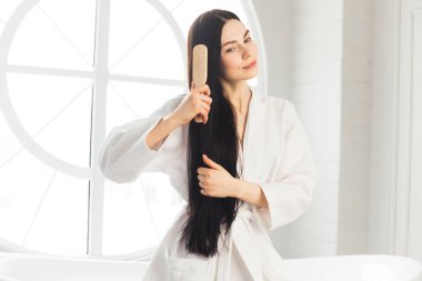 Genç esmer kadın, sağlıklı ve parlak saçlarını tarıyor beyaz bornozla küvette oturuyor.