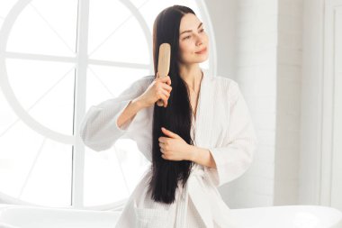 Genç esmer kadın, sağlıklı ve parlak saçlarını tarıyor beyaz bornozla küvette oturuyor.