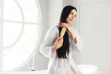 Genç esmer kadın, sağlıklı ve parlak saçlarını tarıyor beyaz bornozla küvette oturuyor.