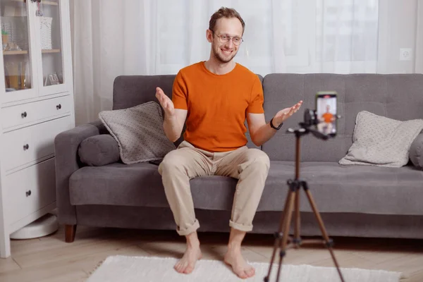 Блогер Hipster man, що записує відеоблог на мобільному телефоні сидить на дивані у вітальні і пояснює і жестикулює руками — стокове фото
