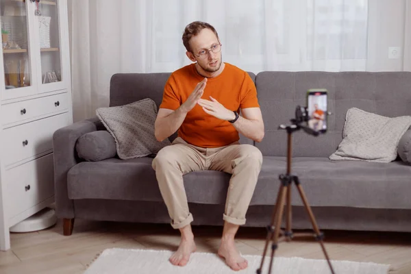 Hipster hombre blogger grabación vlog en el teléfono móvil sentarse en el sofá en la sala de estar y explicar y hacer un gesto con las manos — Foto de Stock