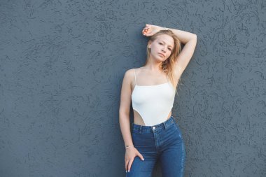 Gri duvar arkasında duran beyaz kolsuz tişörtlü ve mavi kotlu genç sarışın kadın.