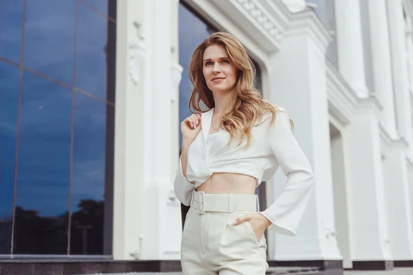 Fashionable woman in white pants and blouse with coat on shoulder posing outdoors — Stock Photo, Image