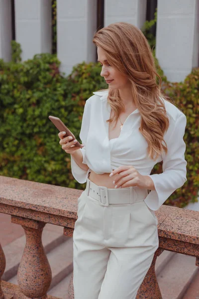 Mujer joven en ropa blanca con teléfono móvil al aire libre, mujer charlando en su teléfono inteligente en la ciudad — Foto de Stock