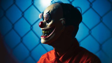 Rahatsız edici palyaço yüzü, geniş gülüşü, şişkin sahte gözleri, hapishane turuncu takımı, arkasında mavi kafes..