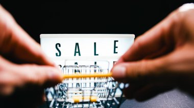 Minyatür bir alışveriş arabasını siyah arka planda aydınlatılmış bir satış tabelasına doğru iten eller. Perakende deneyimi ve alışveriş konsepti.