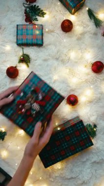 Kırmızı Noel ekose hediye kutuları ve beyaz kürklü bir halıda parlak süsler, arka planda ağaçlar ve ışıklar parlıyor..