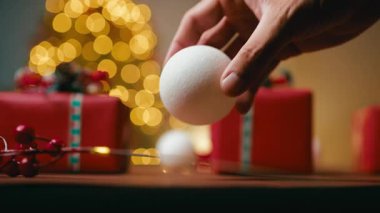 Bir el, kırmızı hediyeler ve altın renkli şenlik arkaplanı olan bir masanın üzerine beyaz bir Noel topu koyar..