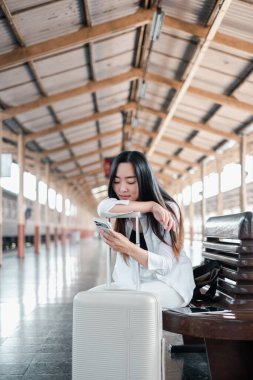 Tren istasyonunda bavullu bir kadın oturuyor, akıllı telefon kullanıyor. Modern seyahat ve ulaşım kavramı.