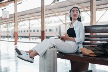 Tren istasyonunda bir bankta oturan genç bir kadın, kulaklık takıyor ve yanında bavuluyla akıllı bir telefon tutuyor..