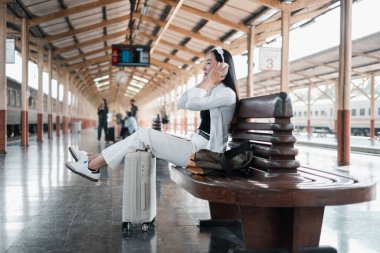 Tren istasyonunda bankta oturan, kulaklıklı müzikten hoşlanan, yanında bavul taşıyan, seyahat macerasına hazır bir kadın..