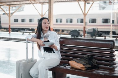 Tren istasyonunda bavul ve tabletle bankta oturan bir kadın, yolculuğa hazır. Modern kentsel ortam.