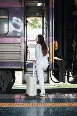 Beyaz elbiseli bir kadın trene biniyor, istasyon peronunda seyahat macerasına hazır..