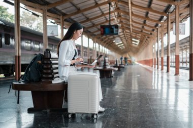 Genç bir kadın bilgisayarını tren istasyonunda kullanıyor. Modern seyahat ve uzak çalışma kültürünü simgeliyor..