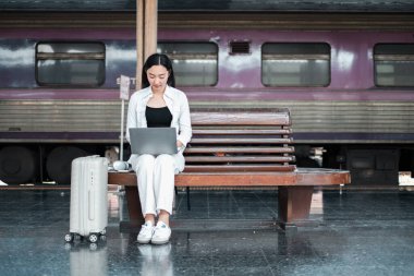 Bir gezgin bir tren istasyonunda bankta oturur, yanında bavullarıyla birlikte bir dizüstü bilgisayarda çalışır, modern seyahat ve teknolojiyi yansıtır..