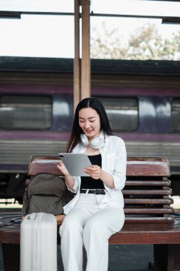 Tren istasyonunda bir bankta oturan bir kadın, bir tablet ve bavulla, seyahat etmeye hazır..