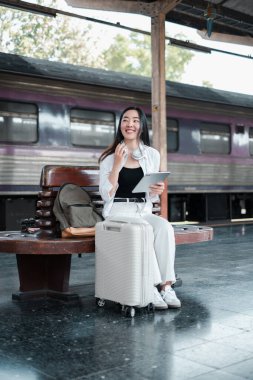 Elinde bavul ve tabletle gülümseyen bir kadın yolcu tren istasyonunda bekliyor, bir sonraki yolculuğu için hazır..