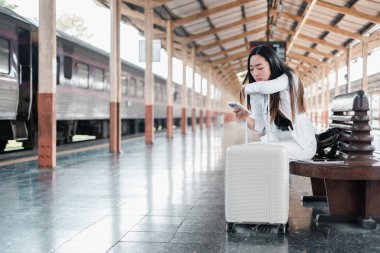 Tren istasyonunda bavuluyla oturan genç bir kadın, akıllı telefonunda seyahat güncellemelerini kontrol ediyor..