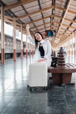 Tren istasyonunda bavullarıyla gülümseyen gezgin, el sallıyor ve yolculuğa hazır. Modern seyahat ve keşif kavramı.