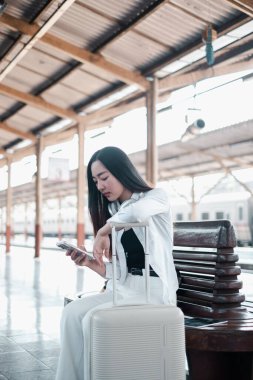 Tren istasyonunda bavuluyla oturan, telefonu kontrol eden, seyahat ve teknolojiyi modern bir ortamda sergileyen bir kadın..