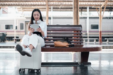 Tren istasyonunda bavulda oturan genç bir kadın, kulaklıklı bir tablet kullanıyor. Seyahat ve teknoloji teması.