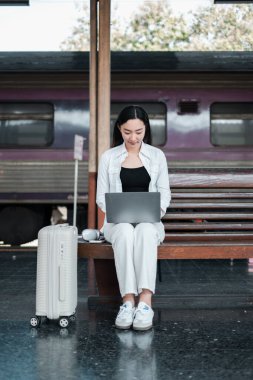 Tren istasyonunda dizüstü bilgisayar kullanan, modern seyahat ve uzaktan çalışma sergileyen sıradan giysili bir kadın..