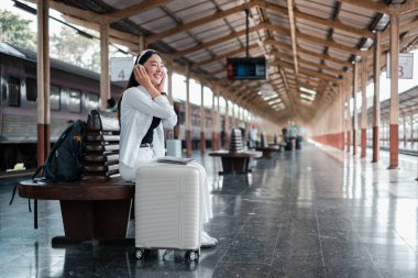 Tren istasyonunda bavulunda oturan, seyahat titreşimlerini kucaklayan, kulaklıklı, gülümseyen bir kadın..