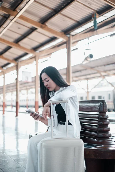 Tren istasyonunda bavuluyla oturan, telefonu kontrol eden, seyahat ve teknolojiyi modern bir ortamda sergileyen bir kadın..