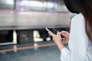 Tren istasyonunda bekleyen bir kadın, arka planda bulanık bir trenle akıllı telefonunu kontrol eder..