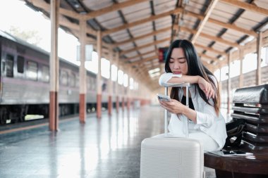 Genç bir kadın tren istasyonunda bir bankta oturuyor, yanında bir bavulla akıllı telefonunu kontrol ediyor, modern seyahati yansıtıyor..