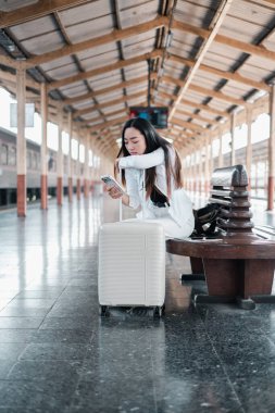 Genç bir kadın tren istasyonunda bavuluyla oturuyor, akıllı telefonunu kullanıyor. Ortam modern ve kentsel..