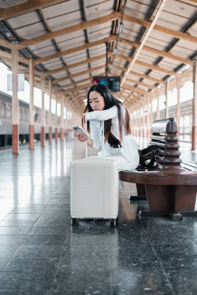 Genç bir kadın tren istasyonunda bavuluyla oturuyor, akıllı telefonunu kullanıyor. Ortam modern ve kentsel..