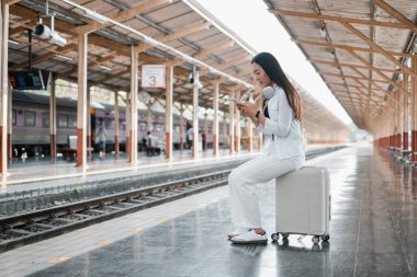 Tren istasyonunda bavulda oturan, akıllı telefon kullanan, kulaklık takan, seyahat etmeye hazır bir kadın..