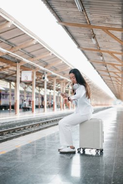 Tren istasyonunda bavulun üzerinde oturan kadın, akıllı bakışları kontrol ediyor, seyahat için hazır..